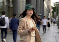 Woman in a beige suit and green cap walking on a city street, holding a matcha cup.