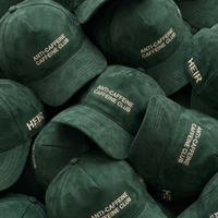 Stack of green caps with 'Anti-Caffeine Caffeine Club' and 'HEIR' text.